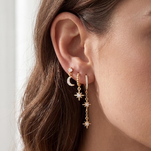 May include: Three gold-toned earrings featuring celestial motifs. One earring has a crescent moon charm with a small stud, another has a star charm on a beaded hoop, and the third is a hoop with a long chain featuring two star charms with crystals.