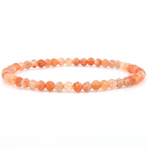 Può includere: Un braccialetto delicato composto da piccole perle di pietra del sole rotonde e sfaccettate. Le perle presentano una tavolozza di colori arancione e pesca traslucido con sottili inclusioni scintillanti, disposte in un cerchio continuo.