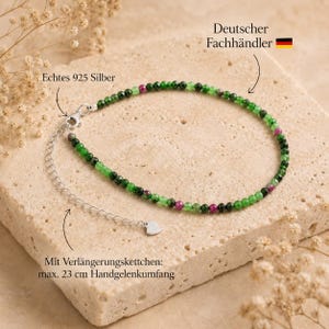 May include: A beaded bracelet featuring small green and magenta gemstone beads with a silver clasp and an adjustable silver chain extension. The text on the image includes 'Echtes 925 Silber', 'Deutscher Fachhändler', and 'Mit Verlängerungskettchen: max. 23 cm Handgelenkumfang'.