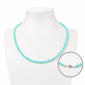 Puede incluir: Un collar de una sola hebra compuesto por pequeñas cuentas redondas y facetadas de color turquesa. El collar cuenta con un cierre de langosta de tono plateado pulido, que se muestra en un detalle circular en la parte inferior derecha.