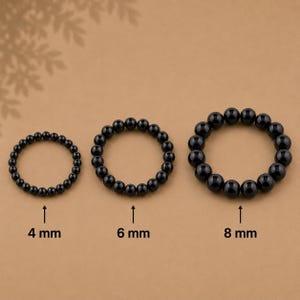 Può includere: Tre braccialetti circolari composti da perline nere lucide, disposti per dimensione. I braccialetti sono etichettati con testo che indica diametri delle perline di 4 mm, 6 mm e 8 mm, con frecce che indicano ogni dimensione corrispondente.