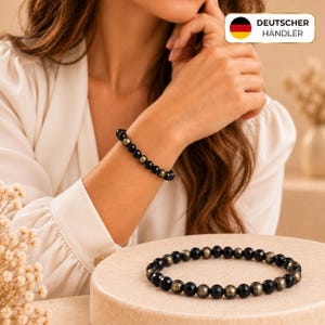 Peut inclure: Un bracelet de perles composé d'une alternance de perles noires facettées et de perles en pyrite aux reflets dorés. Le bracelet est présenté à la fois au poignet et posé sur une surface circulaire, montrant un motif régulier de pierres sombres.