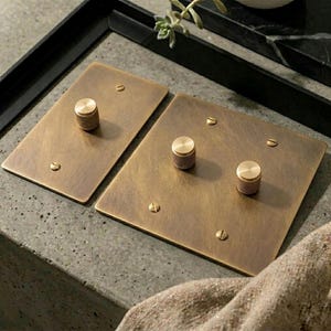 Puede incluir: Dos placas rectangulares de latón para interruptores de luz con acabado cepillado. Una placa tiene un solo mando cilíndrico estriado, mientras que la otra tiene dos mandos a juego. Cada placa está fijada con pequeños tornillos planos.
