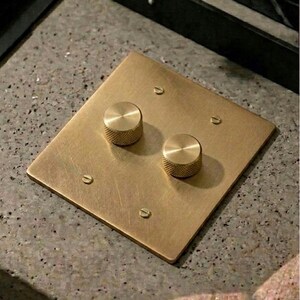 May include: A square, brushed brass light switch plate featuring two circular, knurled control knobs. The plate is secured with four flat-head screws positioned at the corners. The metal surface has a matte, metallic finish with visible texture.