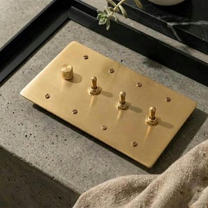 May include: A rectangular brass light switch plate featuring a brushed metallic finish. The plate includes one circular knurled dimmer knob on the left and three vertical toggle switches arranged in a row, all secured by eight small flathead screws.