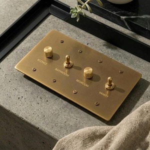 May include: A rectangular brass light switch plate featuring two knurled rotary knobs and two toggle switches. The plate is engraved with the labels SCONCE, BATHROOM, BEDROOM, and VANITY, and is secured with six flat-head screws in a matching brass finish.