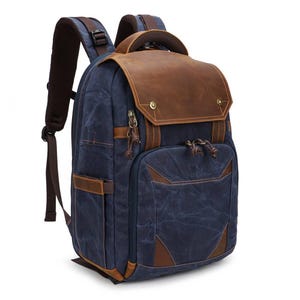 Puede incluir: Una mochila azul marino con una gran solapa superior de cuero marrón y remaches metálicos de color latón. La bolsa incluye correas ajustables para los hombros, un compartimento frontal con cremallera y tiradores de cuero en las esquinas.