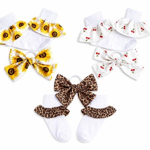 May include: A set of three pairs of white ankle socks with ruffled cuffs and matching hair bows. The patterns include yellow sunflowers, red cherries on a white background, and a brown and black leopard print design on the cuffs and bows.