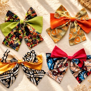 May include: Four hair bows featuring diverse patterns and colors. One bow combines lime green with a multi-colored floral print. Another features orange fabric with a geometric yellow and brown design. A third bow pairs solid yellow with black and white batik patterns. The fourth bow displays a vibrant red, blue, and tan graphic print.