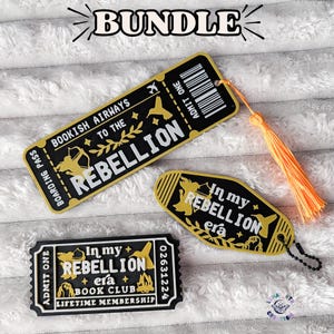 May include: A bundle of three black and gold items: a rectangular bookmark shaped like a boarding pass with a bright orange tassel, a motel-style keychain, and a rectangular ticket-shaped card, all featuring text and graphics related to a rebellion.