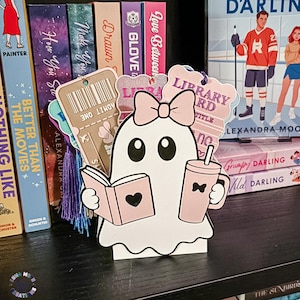 May include: A white ghost-shaped bookmark featuring a pink bow, holding a pink book with a black heart and a pink tumbler with a straw. Behind the ghost are several rectangular bookmarks, including one with a barcode and the text ADMIT ONE.