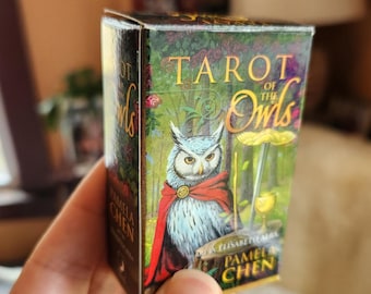 Puede incluir: Una caja de cartas de Tarot of the Owls con un búho azul y gris con una capa roja y una corona dorada. La caja es verde y dorada con el texto "Tarot of the Owls" y "Pamela Chen".