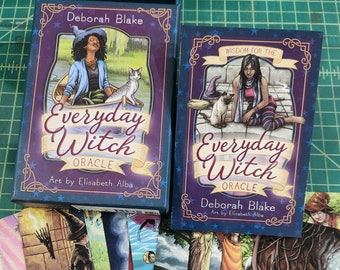 Peut inclure: Un jeu de cartes "Everyday Witch Oracle" de Deborah Blake, avec des illustrations d'Elisabeth Alba. La boîte et les cartes présentent des illustrations de sorcières et de chats. Les cartes sont étalées, affichant des œuvres d'art colorées avec des étiquettes textuelles.