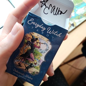 以下が含まれることがあります： 「Everyday Witch」と題された、ネイビーブルーのミニタロットカードボックス。サインが書かれています。箱には、フルートを演奏する魔女のイラスト、音符、黒猫が描かれています。アーティストはデボラ・ブレイクです。