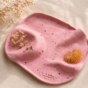 Puede incluir: Una bandeja para joyas rosa de forma irregular con un acabado moteado en morado y blanco. La bandeja presenta hendiduras moldeadas, un accesorio dorado con forma de sol, pequeños pendientes dorados y el texto LARN en relieve.