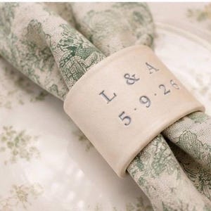 Puede incluir: Un servilletero de cerámica color crema con el texto estampado L & A y la fecha 5.9.26 en tinta gris oscuro. El anillo sostiene una servilleta de tela doblada con un patrón toile verde y blanco, sobre un plato de cerámica a juego.