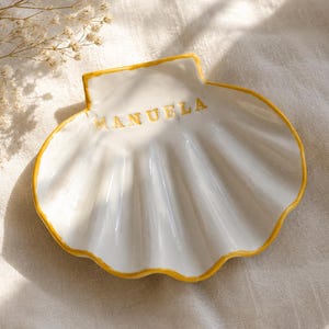 Puede incluir: Un plato decorativo de cerámica blanca con forma de concha, borde pintado en amarillo y el nombre MANUELA grabado en letras amarillas en la parte superior. El plato presenta un borde festoneado y un acabado brillante para objetos.