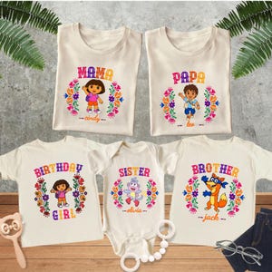 May include: Five off-white shirts and a baby bodysuit featuring colorful Dora the Explorer character graphics. Each garment displays a specific family role label including Mama, Papa, Birthday Girl, Sister, and Brother, along with personalized names.