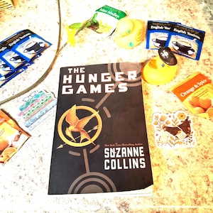 May include: A copy of The Hunger Games by Suzanne Collins with a black cover featuring a gold mockingjay emblem. Surrounding the book are several tea bags, two yellow rubber ducks, one wearing a pirate hat, and two decorative stickers.