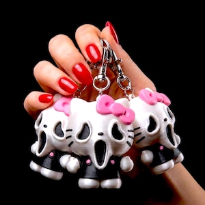 May include: Three white and black character keychains featuring a stylized cat face with a wide open mouth, black eye sockets, and a pink bow on the head. Each figure is attached to a silver-toned metal lobster clasp and split ring connector.