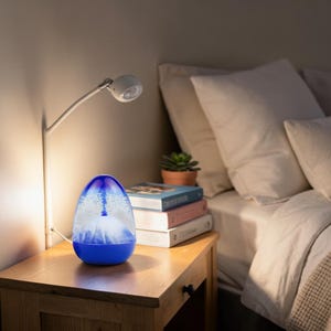 Puede incluir: Una lámpara en forma de lágrima con una base azul y una sección superior transparente que contiene una estructura blanca similar a una montaña sumergida en líquido con pigmentos azules arremolinados, emitiendo un brillo suave.