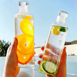 May include: Two clear glass water bottles with narrow necks and transparent caps. One bottle contains water infused with bright orange slices, while the other bottle features cucumber slices and printed time markers for hydration tracking.