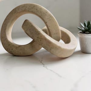 Könnte beinhalten: Eine beige dekorative Kettengliedskulptur mit gesprenkelter Textur, bestehend aus zwei ineinandergreifenden ovalen Schlaufen. Das Objekt hat eine glatte, matte Oberfläche und steht neben einer kleinen grünen Sukkulente in einem Topf.