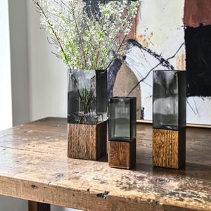 May include: Three rectangular vases featuring a combination of dark gray translucent glass tops and textured brown wooden bases. The tallest vase on the left contains thin branches with small white flowers, while the other two remain empty.