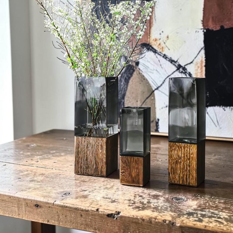 May include: Three rectangular vases featuring a combination of dark gray translucent glass tops and textured brown wooden bases. The tallest vase on the left contains thin branches with small white flowers, while the other two remain empty.