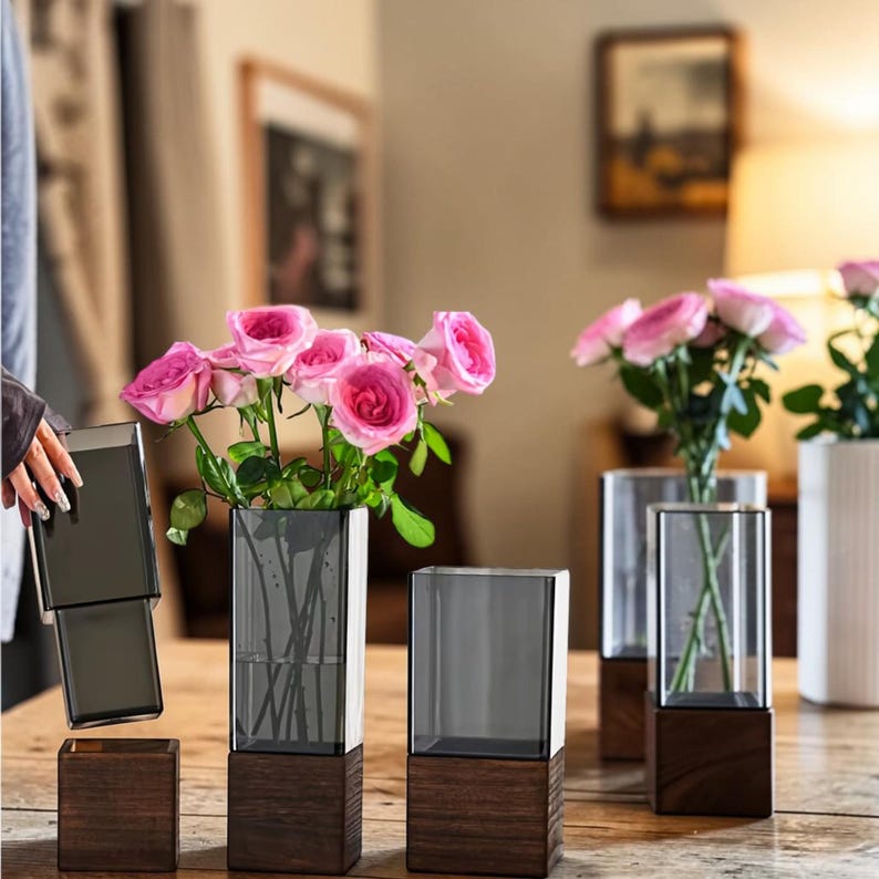 May include: A set of rectangular vases featuring dark gray glass tops and solid dark wood bases. Some vases are empty, while others contain pink roses with green stems. The design consists of a clear glass sleeve that fits into a wooden block base.