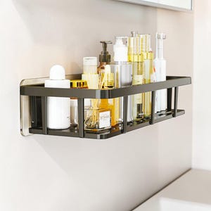 Può includere: Una mensola da bagno rettangolare in metallo nero montata a parete, contenente vari articoli da toeletta tra cui flaconi bianchi, contenitori in vetro trasparente con dettagli dorati, una piccola bottiglia di vetro con etichetta WHITE e uno spazzolino elettrico.