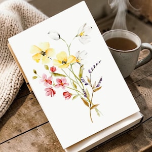 Könnte beinhalten: Eine weiße Grußkarte mit einer Aquarellillustration eines Wildblumenstraußes. Der Strauß besteht aus gelben, rosa, weißen und violetten Blüten mit grünen Stielen und Blättern. Die Karte liegt auf einer Holzoberfläche neben einer Tasse.