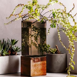 May include: A tall, square-shaped glass vase featuring a two-tone design with a translucent, smoky-grey upper section and a reflective, metallic copper-colored base, containing delicate branches with small white flowers.