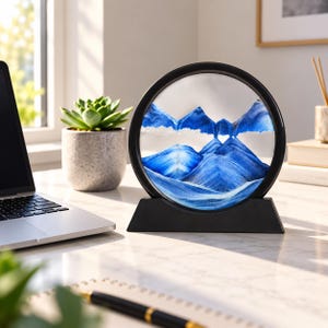 Peut inclure: Un objet d'art en sable circulaire présentant des couches de sable bleu et blanc formant des sommets montagneux à l'intérieur d'un cadre noir. L'ensemble est monté sur une base triangulaire noire solide, créant un paysage visuel.