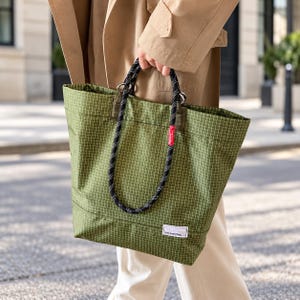 May include: A green grid-patterned tote bag featuring black and white rope handles, silver metal rings, and a small white rectangular label with the text tote brand bag. A small red fabric tag is attached to one of the black rope handles.