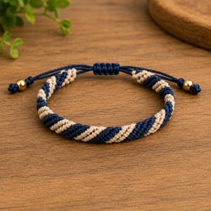 May include: A braided cord bracelet featuring a repeating spiral pattern of navy blue and beige threads. The bracelet includes an adjustable sliding knot closure and two gold-toned metallic beads at the ends of the pull strings.