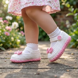 May include: A pair of white crocheted baby shoes with pink trim and a small pink bow on the toe. The shoes feature a strap across the top secured with a pink button. The shoes are worn with white socks and a floral patterned dress is visible above.
