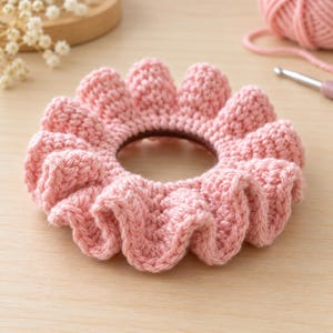 May include: A light pink crocheted hair scrunchie with a ruffled, textured edge. The circular accessory features a dense, hand-stitched yarn construction with a dark brown elastic band visible in the center opening of the fabric.