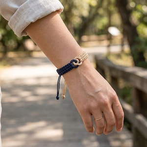 May include: A two-tone cord bracelet featuring a dark navy blue section and a light beige section connected by a central knot. The bracelet has a woven texture with two loose cord ends hanging down, showcasing a simple and functional design.