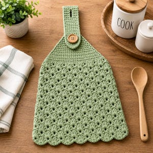 May include: A sage green crocheted hanging kitchen towel featuring a textured shell stitch pattern, a solid crocheted top section, and a single round wooden button closure at the top tab for securing the towel to a handle or rail.