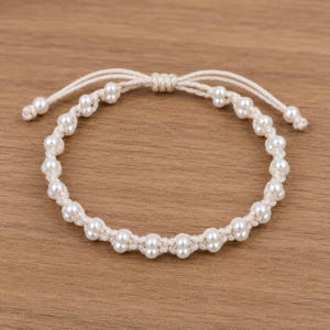 May include: A bracelet featuring a series of small, round, white beads woven into a light beige cord. The design includes a central circular band of beads with two adjustable cord ends extending from a knotted center point at the top of the piece.