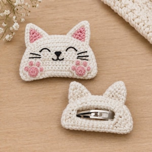 May include: Two off-white crochet cat hair clips featuring pink inner ears and paw pads. The top clip displays a stitched black face with eyes, nose, and whiskers. The bottom clip shows the reverse side with a silver metal snap hair clip attached.