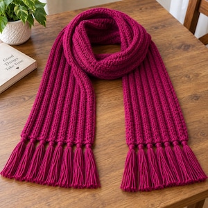 May include: A vibrant magenta knitted scarf featuring a prominent vertical ribbed texture throughout the length. The scarf ends are finished with a series of matching magenta yarn tassels, creating a uniform and textured appearance.
