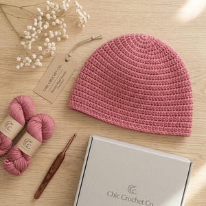 Puede incluir: Un gorro de ganchillo color rosa empolvado, dos ovillos de lana a juego, un ganchillo de madera y una etiqueta de marca Chic Crochet Co. Merino Wool Beanie. Los artículos se disponen junto a una caja de regalo blanca y paniculata.