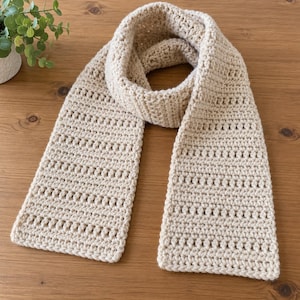 May include: A beige crochet scarf featuring a repeating pattern of horizontal rows with textured stitches and openwork gaps. The scarf is constructed from yarn and displays a consistent light tan color throughout its entire length.