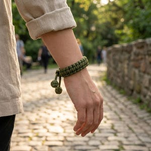 Peut inclure: Un bracelet en paracorde vert olive présentant un motif de nœuds tressés épais et deux breloques sphériques nouées pendantes à l'extrémité. Le bracelet est fabriqué à partir d'un cordon durable et s'enroule autour du poignet avec une finition texturée.