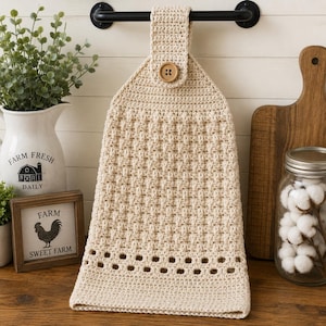 May include: A beige crochet hanging kitchen towel featuring a basketweave texture, a single wooden button closure at the top loop, and a decorative openwork stitch pattern near the bottom edge, suspended from a black metal towel bar.