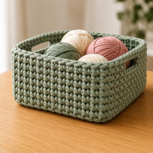 May include: A rectangular sage green crochet basket with integrated side handles, containing four round balls of yarn in shades of cream, sage green, and dusty rose. The basket features a thick, textured stitch pattern throughout its structure.