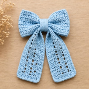 May include: A light blue crochet bow hair accessory featuring a central knot and two long, dangling tails. The bow is constructed with a dense stitch pattern on the top loops and a decorative openwork, grid-like pattern on the two hanging tails.