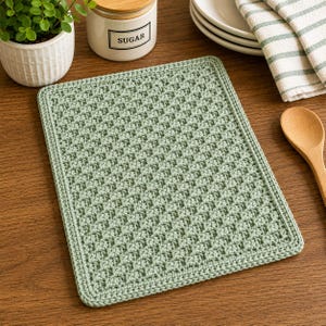Puede incluir: Una alfombrilla rectangular de ganchillo en color verde salvia que presenta un patrón texturizado de punto de gofre con un borde sólido. La alfombrilla está hecha de hilo y muestra un diseño de cuadrícula repetitivo y consistente en toda su superficie.
